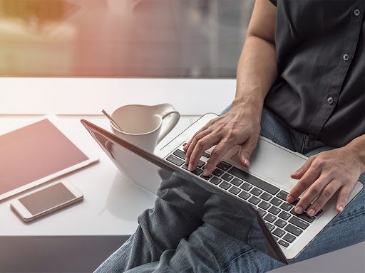 person sitting on a bench outside with coffee, working on their Apple laptop